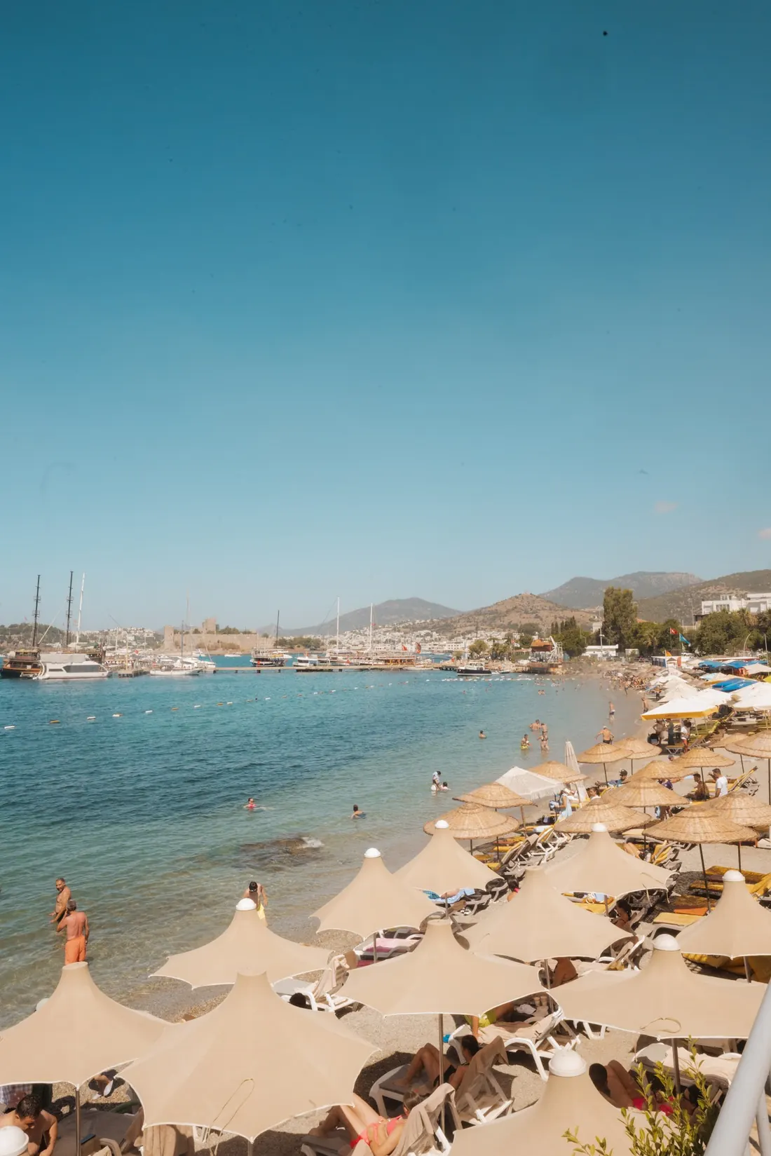 A crowded coastal beach of straw umbrellas below a harbour.