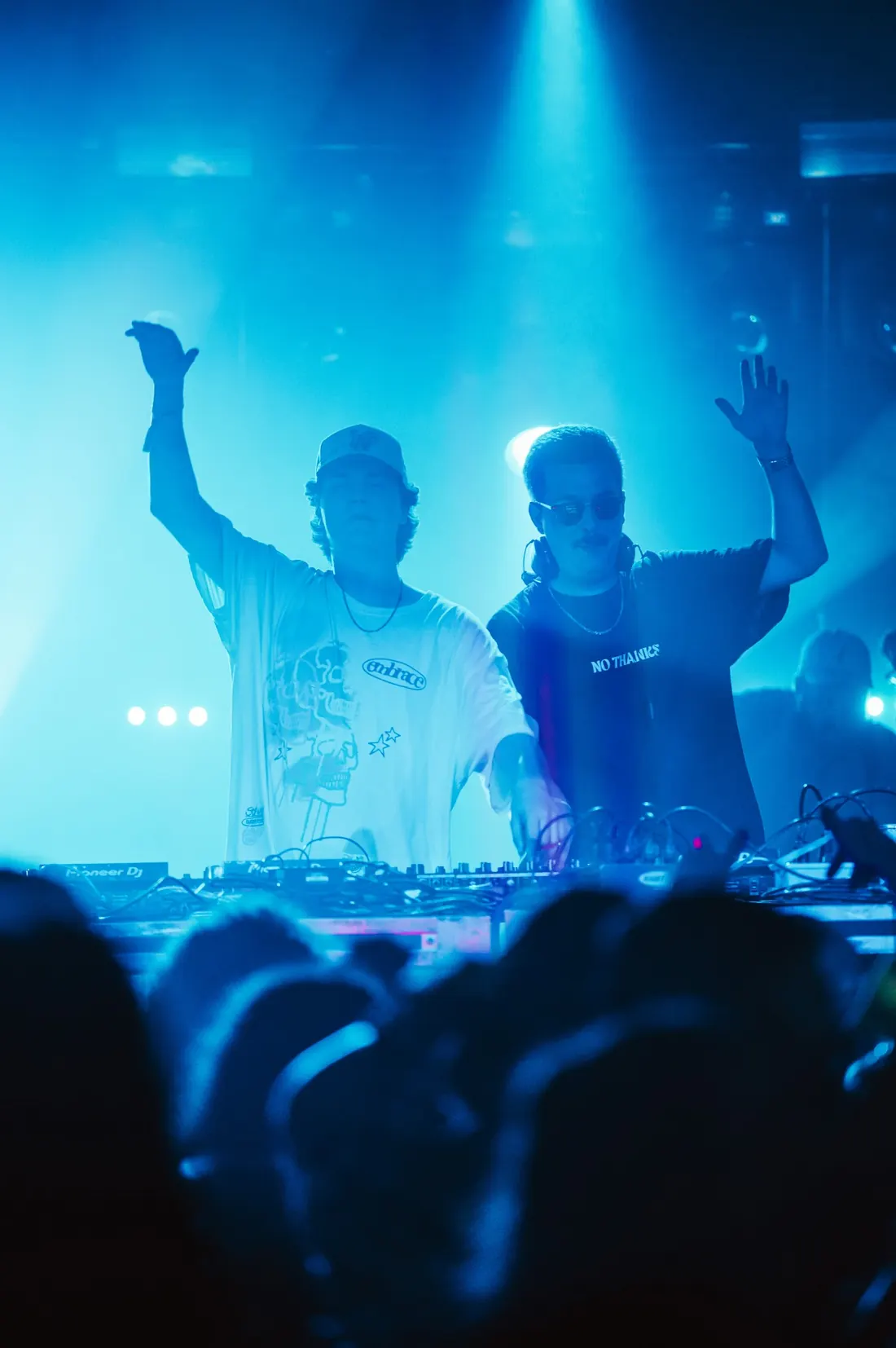 Two DJs with arms raised in blue stage light above a crowd.
