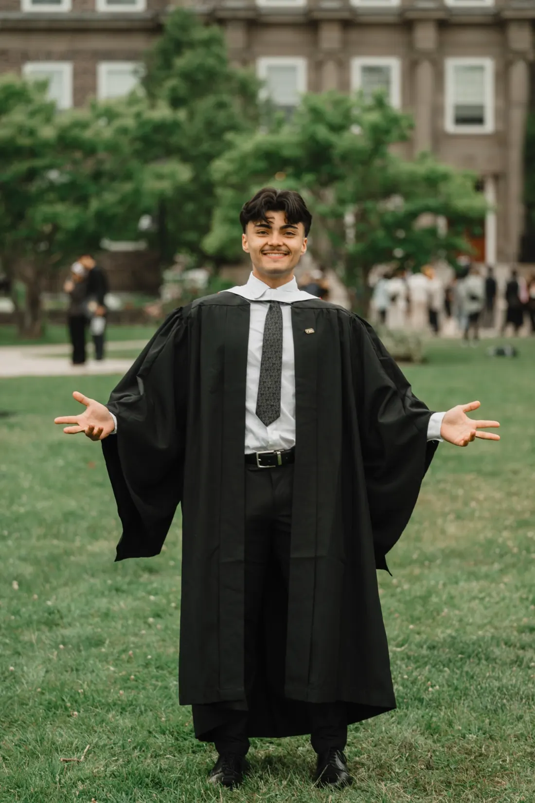 A graduate in cap and gown on a campus lawn with arms open.