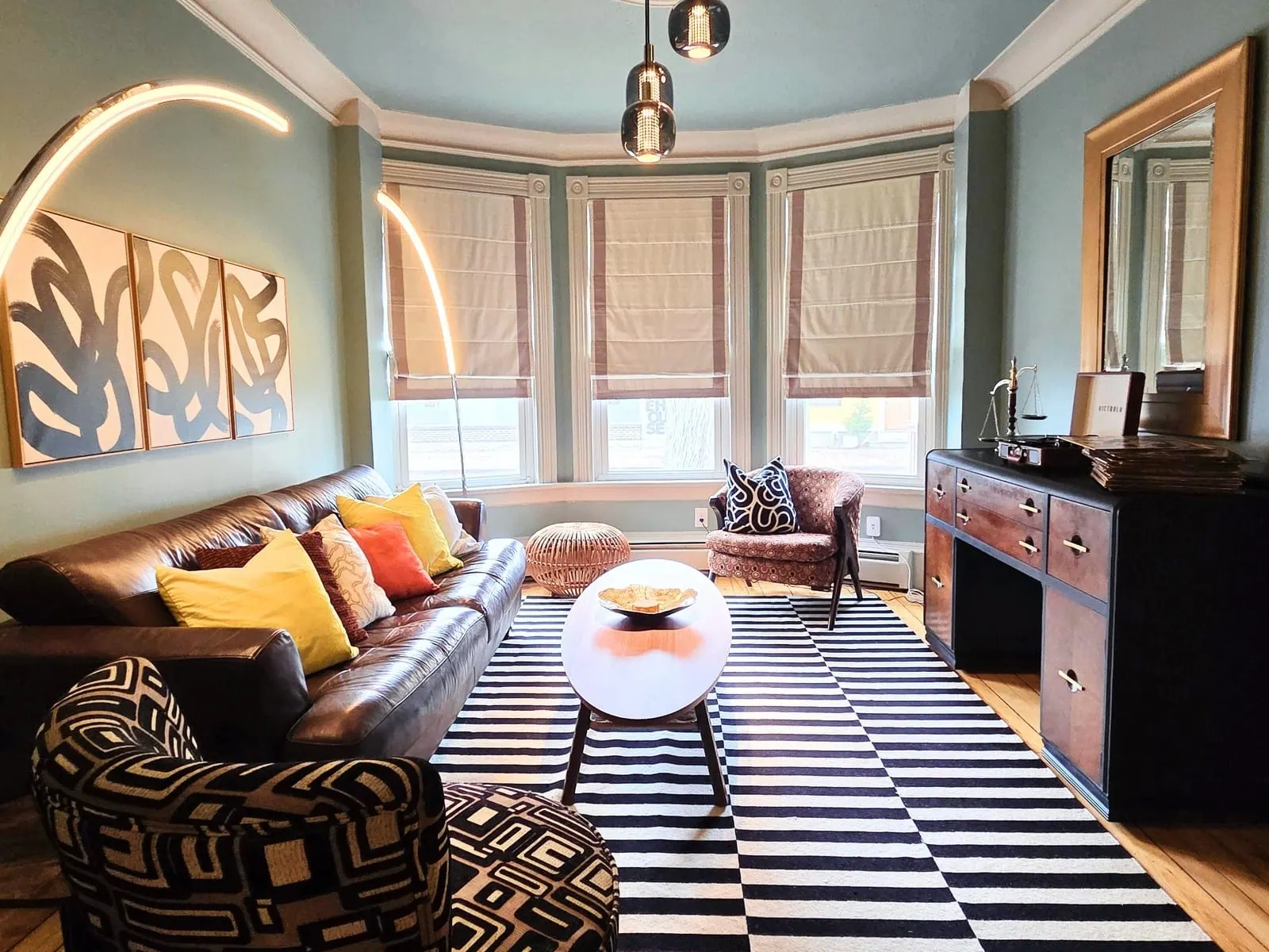 Living room with a black-and-white striped rug, leather sofa with mustard and orange cushions, and bay windows with roman shades