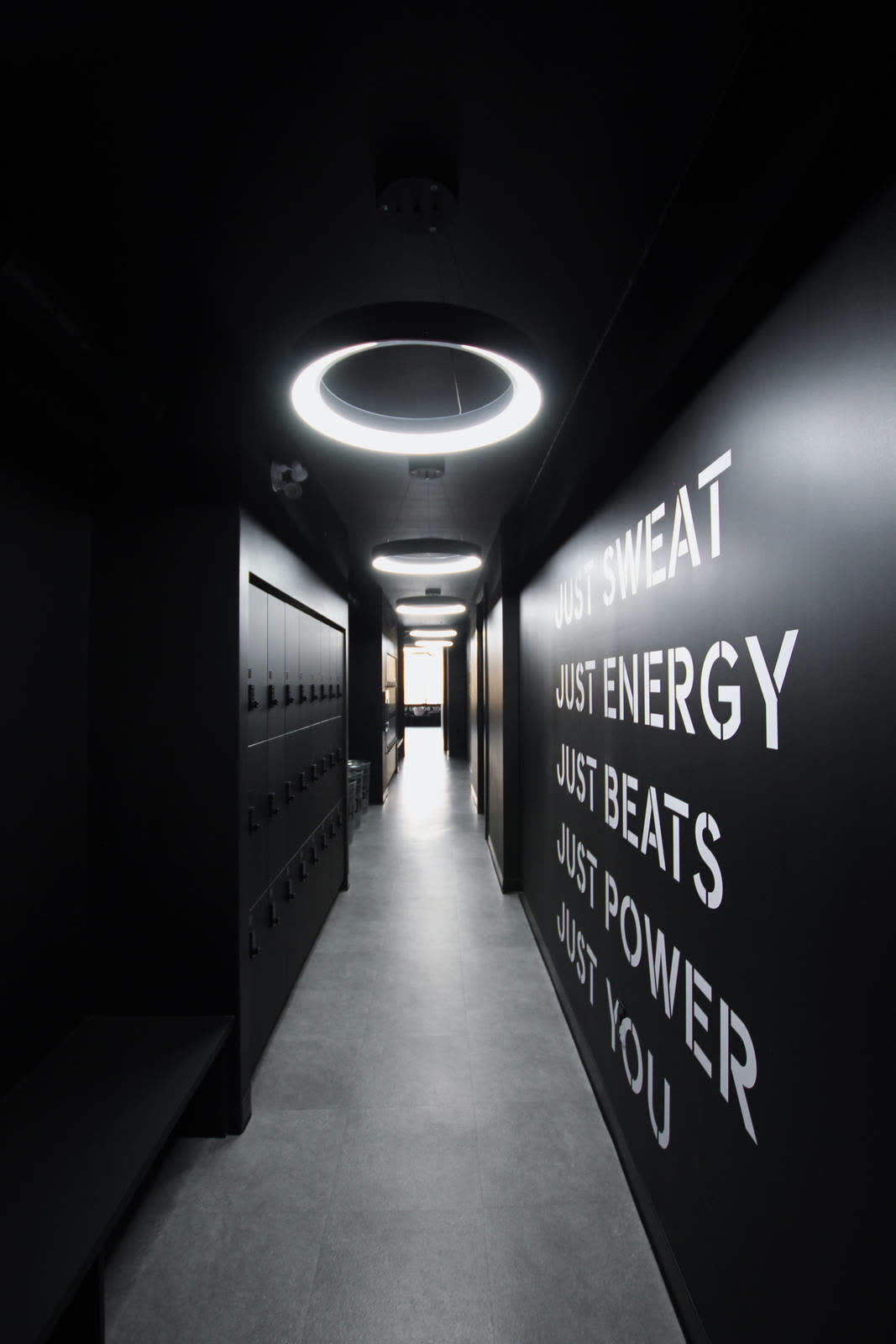Locker corridor under ring lights; the wall reads JUST SWEAT, JUST ENERGY, JUST BEATS, JUST POWER, JUST YOU